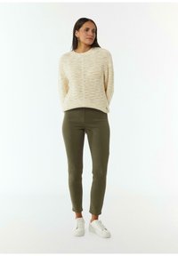 Übergroßer cremefarbener Pullover mit strukturiertem Muster, kombiniert mit eng anliegenden olivgrünen Hosen und weißen Sneakern, vor einem schlichten Hintergrund stehend.