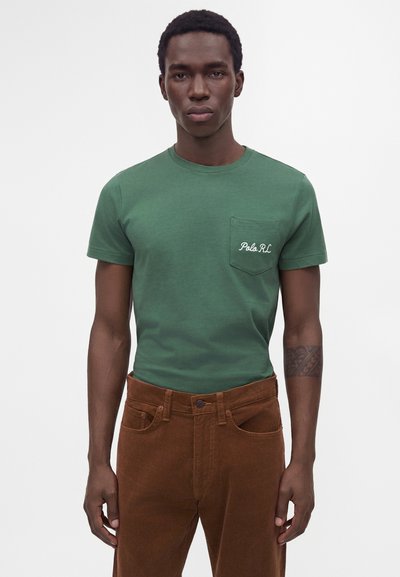 Uomo che indossa una maglietta Polo RL verde con taschino sul petto e pantaloni marroni, in piedi davanti a uno sfondo bianco uniforme.