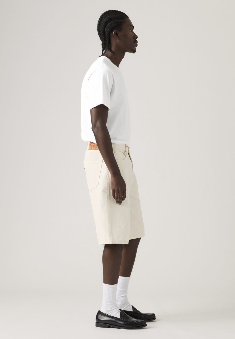 Homme aux cheveux tressés debout de profil, portant un t-shirt blanc, un short beige jusqu'aux genoux, des chaussettes blanches et des mocassins noirs, sur un fond uni.