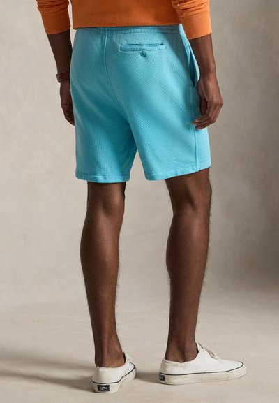Homme portant un short bleu clair et des baskets blanches, debout avec une main posée le long du corps sur un fond uni.