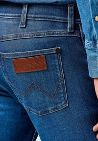 Donkerblauwe denim jeans met een bruine leren Wrangler-label. Heeft een herkenbaar stikselontwerp op de achterzak en metalen hardwareaccenten.