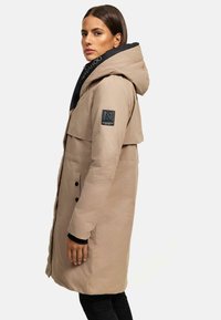 Manteau long beige avec une capuche noire. Dispose d'un col distinctif, de boutons sur le devant et d'une patch de marque sur la manche. Texture lisse.