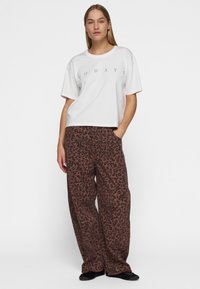 Donna in piedi con le mani in tasca, indossa una t-shirt bianca "10 DAYS" e pantaloni larghi a stampa leopardata marrone con scarpe nere.