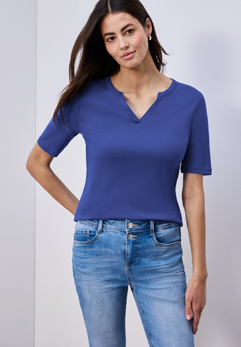 Femme aux longs cheveux noirs portant un haut bleu à manches courtes et un jean bleu clair, debout avec une main sur la hanche.