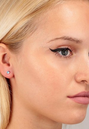 Orecchino a bottone in argento con una pietra preziosa rotonda di colore azzurro chiaro. Primo piano di un volto con eyeliner audace, pelle liscia e un colore di labbra neutro.