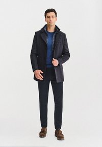 Męski ciemnoszary płaszcz w kratę z podwójnym zapięciem, założony na niebieski sweter, zestawiony z ciemnymi spodniami i brązowymi butami. Prosty, nowoczesny wygląd.