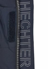 Marineblauwe jas met een glanzende rits. "HECHTER" is in witte stippen langs de voorkant gedrukt, met een gestructureerde zwarte klittenbandpatch.