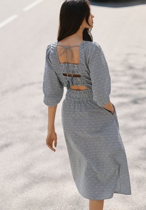 Žena hoda ulicom noseći sivo-bijelu midi haljinu s prugama, naboranim rukavima i otvorenim detaljima na leđima vezanim vrpcama.