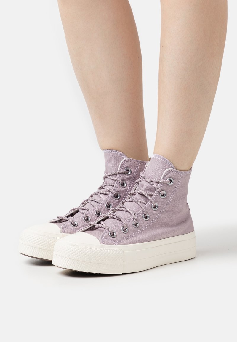 Paire de baskets montantes en toile violet clair avec semelles et lacets en caoutchouc blanc, portées pieds nus sur un fond uni.