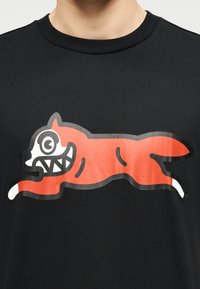 Μαύρο βαμβακερό μπλουζάκι με γραφικό σχέδιο ενός τρέχοντος πορτοκαλί πλάσματος με λευκό πρόσωπο και μαύρες λεπτομέρειες, στο μπροστινό μέρος.