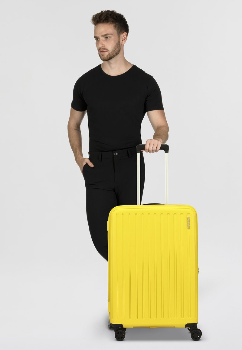 Valigia rigida gialla brillante con righe verticali, manico telescopico e quattro ruote, posizionata accanto a una persona in abbigliamento nero.