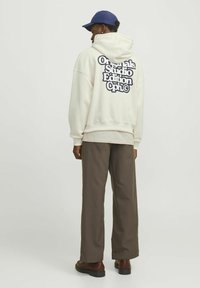 Felpa crema con grande testo grafico in navy sulla schiena, abbinata a pantaloni larghi marroni e scarpe marroni, indossata con un berretto blu.