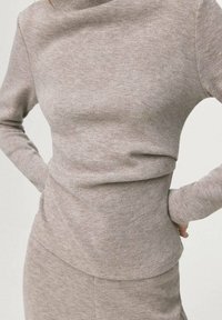 Haut en maille beige clair avec un col montant et des manches longues, présentant une coupe ajustée et un léger drapé à la taille.