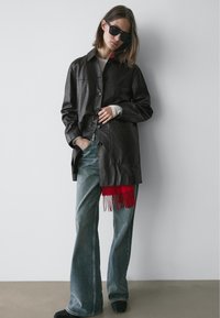 Zwarte leren overhemdjas met een gestructureerd oppervlak, knopen aan de voorkant, gecombineerd met lichtblauwe uitlopende jeans en een rode franjerand.