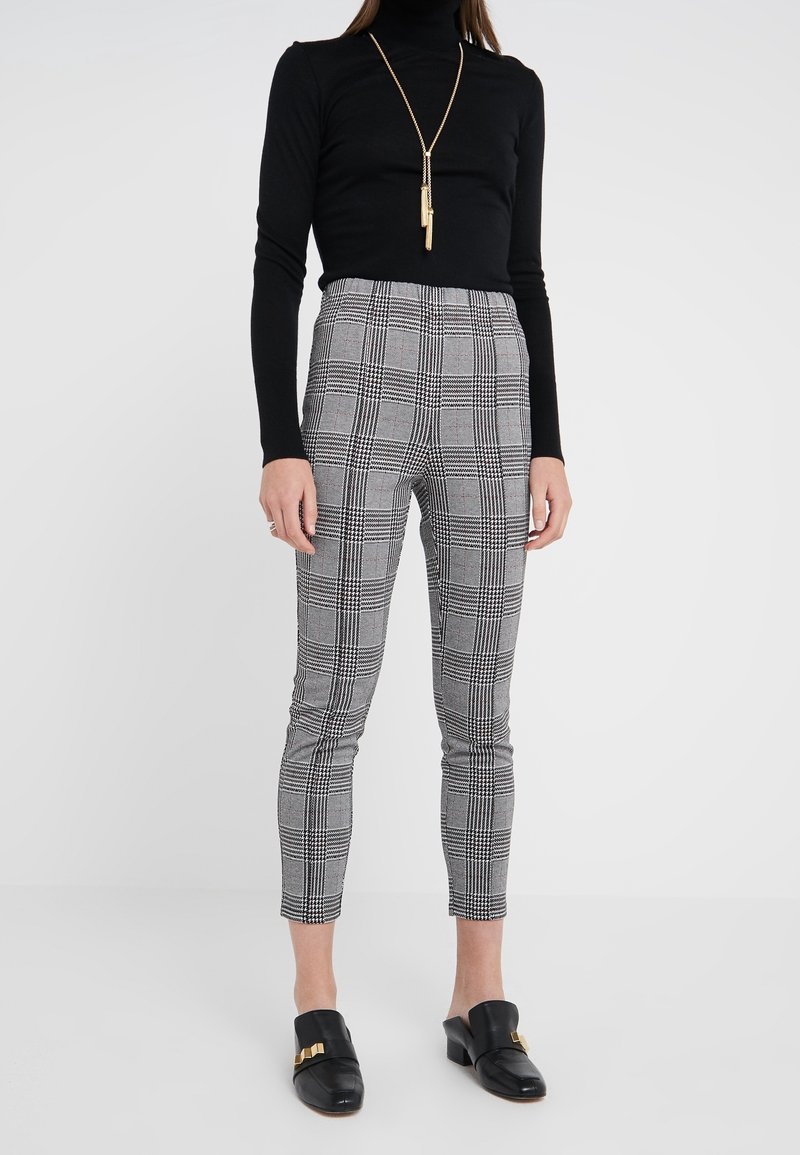 Maglione a collo alto nero abbinato a pantaloni a righe grigio tartan con linee verticali e orizzontali, e scarpe nere con un accento dorato.