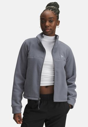 Giacca in pile grigia con zip e colletto alto, caratterizzata da un design aderente e due tasche frontali, indossata sopra una maglietta bianca e pantaloni neri.