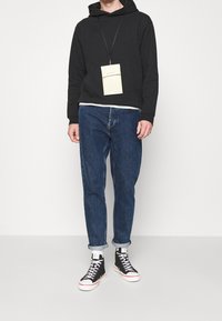 Sudadera negra con capucha y bolsillo tipo canguro, combinada con vaqueros azules. Zapatillas negras de caña alta con suelas blancas. Una etiqueta de color neutro cuelga del cuello.