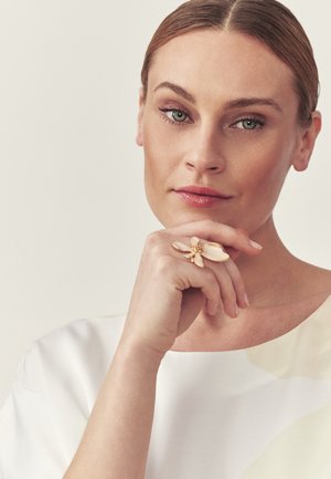 Heledan nahaga ja roheliste silmadega naine, valge toppi kandmas, lõua toetamine käele, mis on kaunistatud suure kuldse lillekujuga sõrmusega.