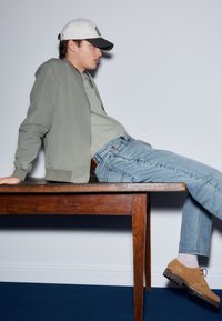 Grøn bomberjakke, grå t-shirt, blå jeans, beige ruskindssko. Model siddende på et træbord foran en ensfarvet baggrund.