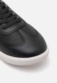 Zwarte leren sneaker met een gestructureerde bovenkant, ronde neus en witte rubberen zool met een ruitpatroon. Veters zijn door oogjes geregen.