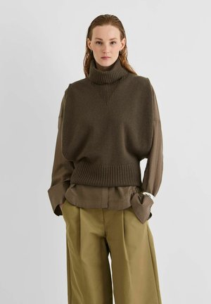 Femme aux cheveux mouillés portant un pull sans manches à col roulé marron foncé sur une chemise à manches longues et un pantalon beige à jambes larges, debout les mains dans les poches.