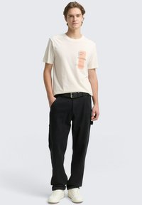 Cremfarbendes Baumwoll-T-Shirt mit Grafikdruck, kombiniert mit schwarzen, locker sitzenden Hosen und weißen Sneakers. Einfaches Gürteldetail sichtbar.