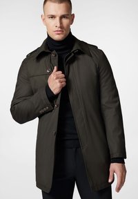 Ciemnozielony płaszcz trench z wyraźnym kołnierzem, z przednimi guzikami i kieszeniami bocznymi. Noszony na czarnym golfie dla kontrastu. Gładki materiał.