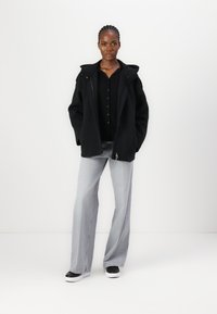 Black zip-up coat with a hood, layered over a black button-up shirt, paired with light gray wide-leg jeans and black and white shoes.