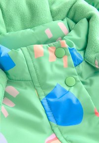 Groene fleece-gevoerde jas met een gladde buitenlaag, versierd met kleurrijke geometrische patronen in blauw en roze, afgewerkt met groene drukknoopjes.