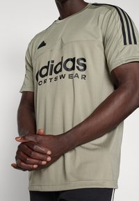 Ljus olivgrön Adidas sport-t-shirt med svarta ränder på ärmarna, med vertikal ribbad textur och en stor svart logotyp på framsidan.