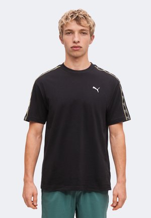 Młody mężczyzna z krótkimi kręconymi blond włosami, ubrany w czarną koszulkę Puma z logo i paskami na rękawach, stojący na jasnym tle.