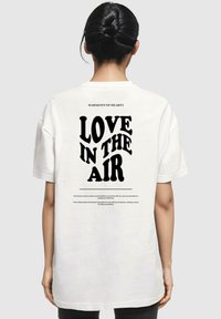 Witte katoenen T-shirt met op de achterkant bold zwarte tekst die leest "LIEFDE IN DE LUCHT", vergezeld van decoratieve tekst boven en onder.