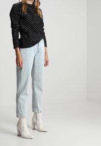 Zwarte lange mouwen top met diagonale zilveren strepen, gecombineerd met lichtblauwe high-waisted jeans en witte enkellaarsjes met dikke hakken.