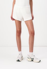 White ribbed shorts with an elastic waistband, featuring a relaxed fit and rounded hem, paired with white and green sneakers and white socks.