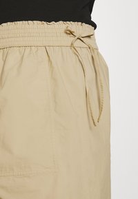 Beige hame, jossa on kootut elastiset vyötärönauhat ja nyöri, sekä koristeellinen sivutasku, valmistettu kevyestä kankaasta.