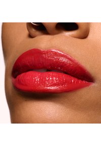 Heldere rode lippen met een glanzende afwerking, met een uitgesproken vorm en textuur. De huidskleur is warm, wat een contrasterend effect creëert.