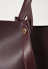 Sac à main en cuir bordeaux avec une surface texturée, des poignées courbées et des accessoires en or. La vue rapprochée met en valeur les coutures et la structure du sac.