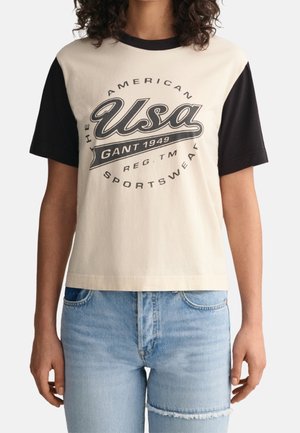 Personne portant un t-shirt beige et noir à manches courtes avec le texte "USA GANT 1949 American Sportswear" et un jean bleu clair déchiré.