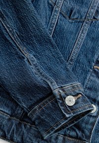 Denim jacket cuff featuring blue fabric, visible stitching, and a metal button with engraved brand name. Close-up of textured material.