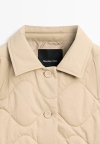 Beżowa pikowana kurtka z rozłożystym kołnierzem, zapięciem na zatrzaski i gładką fakturą. Etykieta Massimo Dutti widoczna wewnątrz kołnierza.