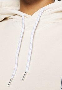 Béžová mikina s kapucí a šňůrkou s modro-bílým pruhovaným vzorem. Tkanina je měkká, s žebrovanými detaily kolem výstřihu.