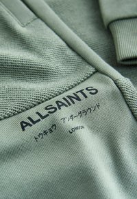 Tejido de punto verde con puños acanalados, con texto de marca en negro. Las costuras y la textura visibles aportan detalle; incluye caracteres japoneses.