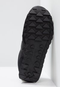Semelle de chaussure noire avec des crampons triangulaires surélevés pour une meilleure traction, inclinée contre un mur et un sol blancs.