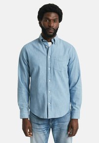 Lyseblå skjorte med knapper i blødt stof, der har en brystlomme og lange ærmer. Kombineret med mørkeblå denimjeans.