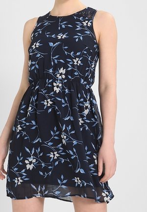 Mouwloze marineblauwe jurk met blauw-witte bloemenprint, elastische taille en doorschijnende zoom gedragen door een persoon tegen een effen achtergrond.