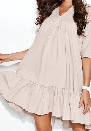 Kvinde iført løs, let beige knælængde kjole med flæsekant, korte ærmer og V-hals, iført hvidt ur og guldkæde.