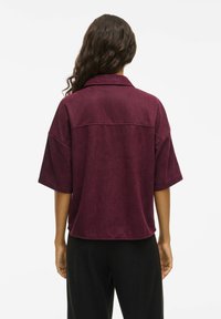 Bordó krāsas pārizmēra krekls ar apkakli, īsām piedurknēm un gludu faktūru. Tam ir aizmugurējā šuve un viegli izliektas apakšmala.