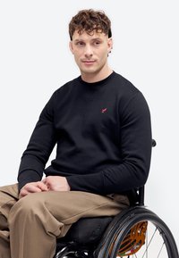 Homme aux cheveux bouclés portant un sweat-shirt noir et un pantalon beige assis dans un fauteuil roulant sur un fond uni.