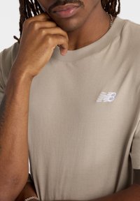 Beige bavlněné tričko s kulatým výstřihem, na hrudi s bílou vyšívanou logem New Balance, hladká textura a volný střih.