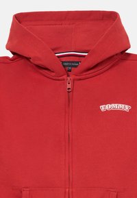 Κόκκινη ζακέτα με φερμουάρ και κουκούλα, μπροστινές τσέπες και την επιγραφή "TOMMY" τυπωμένη στα λευκά στην επάνω αριστερή πλευρά του στήθους.